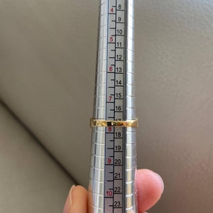 May include: A gold ring being measured on a metal ring sizer. The ring sizer has markings from 5 to 26.