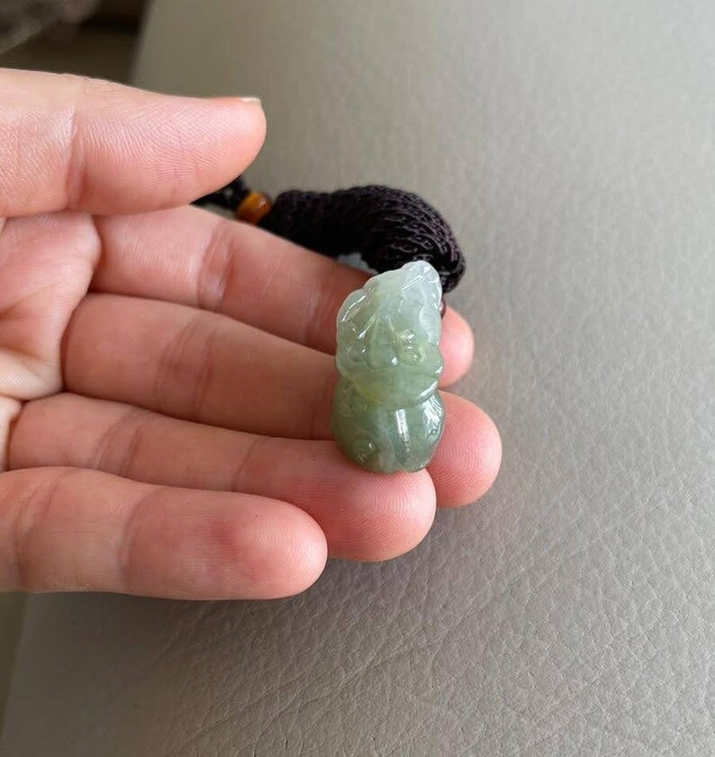May include: A green jade pendant carved in the shape of a mythical creature. The pendant is attached to a brown cord.