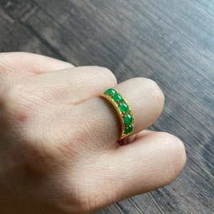 May include: A gold ring with five green oval-shaped gemstones set in a row. The ring is on a person's finger.
