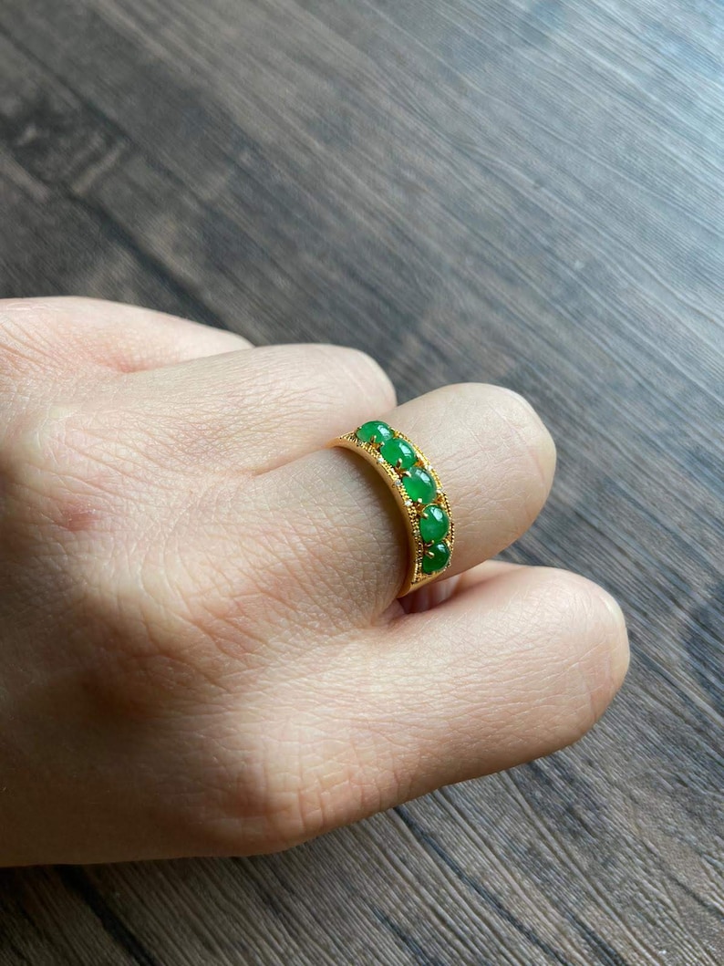 May include: A gold ring with five green oval-shaped gemstones set in a row. The ring is on a person's finger.