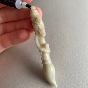 May include: A white jade pendant carved in the shape of a dragon holding a pearl. The pendant is attached to a black cord with white and green beads.