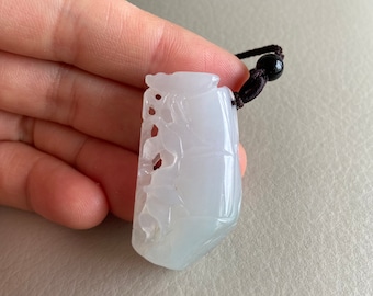 May include: A white jade pendant carved with intricate designs, hanging from a brown cord with a black bead.