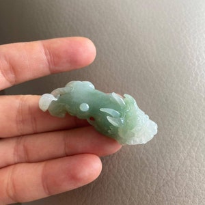 May include: A jade green dragon pendant carved from jade. The dragon is in a curled position with its head facing to the right. The pendant is smooth and polished.