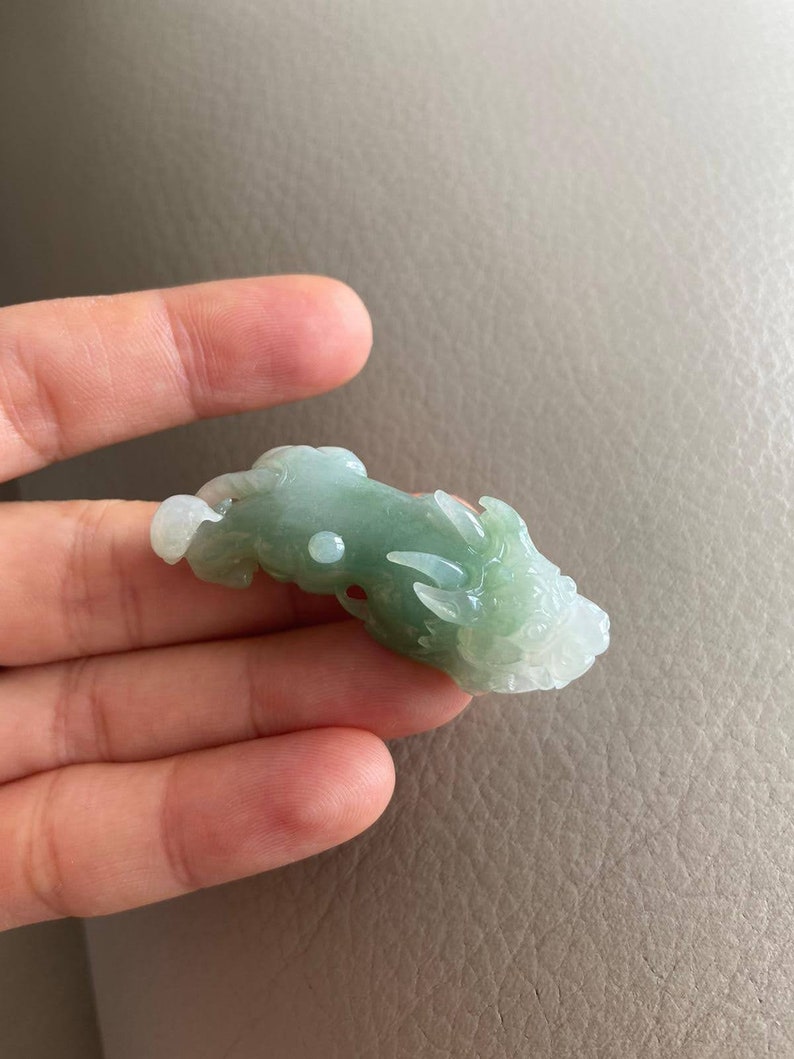 May include: A jade green dragon pendant carved from jade. The dragon is in a curled position with its head facing to the right. The pendant is smooth and polished.