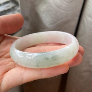 May include: A jade bangle bracelet with a pale green and white color scheme. The smooth, polished surface of the bracelet reflects light, highlighting the natural variations in the stone. This elegant piece of jewelry is a classic accessory.