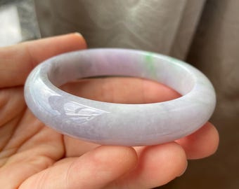 May include: A polished jade bangle bracelet, displaying a mix of lavender, white, and green hues. The smooth, circular design reflects light, showcasing the natural patterns within the stone. This bracelet is a classic piece of jewellery.