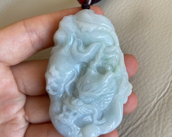 May include: A jade pendant carved with a detailed dragon design. The pendant is a light green color and has a smooth, polished finish. It is attached to a brown cord.