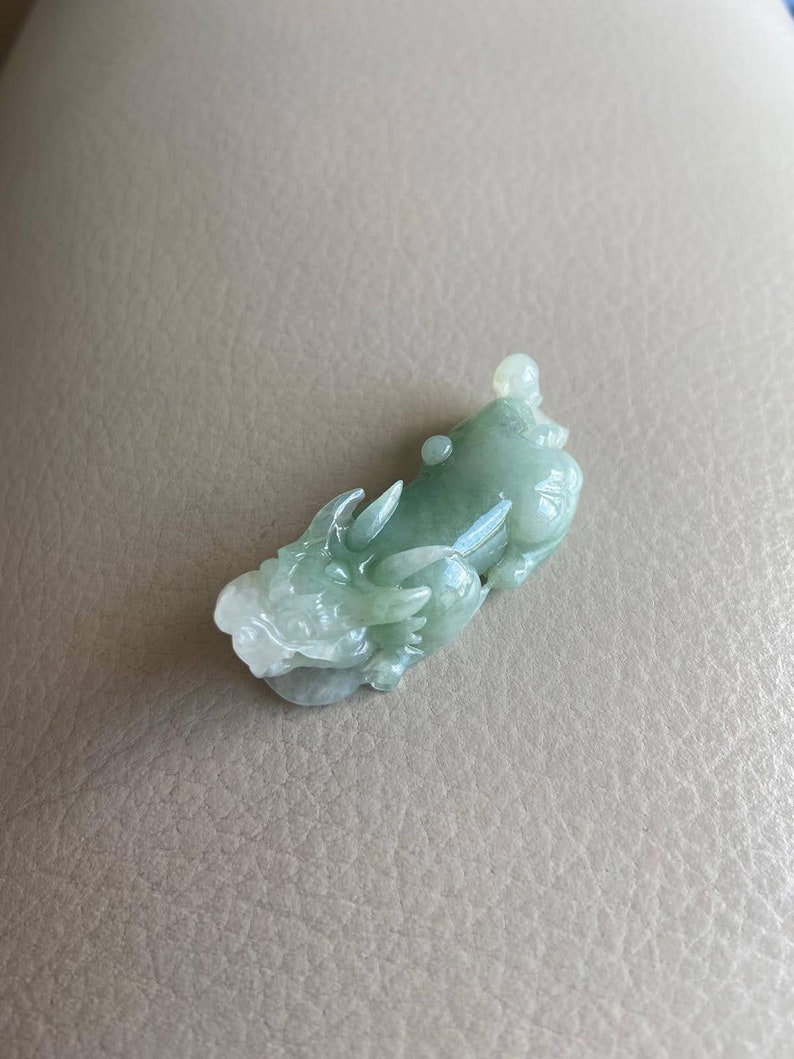 May include: A jade green dragon pendant carved from jade. The dragon is in a curled position with its head raised and its tail curled around its body.