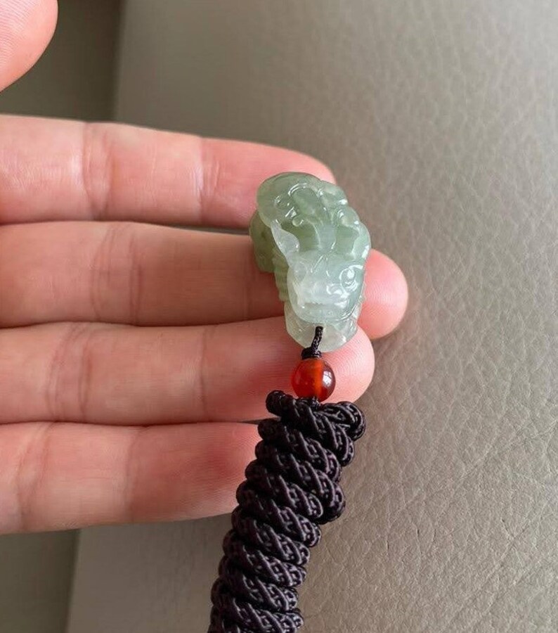 May include: A green jade pendant carved with a detailed design, attached to a brown cord with a red bead.