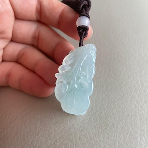 May include: A light green jade pendant carved in the shape of a leaf with intricate details. The pendant is attached to a brown cord with a white bead.