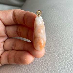 May include: A pendant with a gold-coloured bail and a smooth, elongated jade stone. The jade transitions from white to orange, with a polished finish. The pendant is held in a hand.
