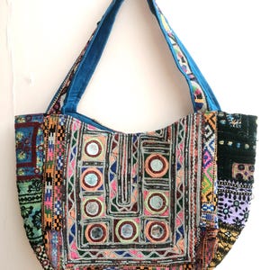 Puede incluir: Un bolso tote grande y colorido con un diseño de patchwork. El bolso presenta una variedad de telas en colores vibrantes, adornadas con espejos y bordados intrincados. Las asas son de color azul liso y el bolso cuelga sobre un fondo blanco.