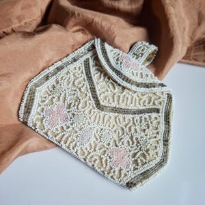 May include: Vintage ivory beaded purse with floral patterns and a small handle. The purse has a flap closure and is trimmed with silver beads. Pink and light green floral accents are visible on the front. The purse is set against a peach-coloured fabric.