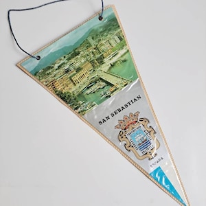 May include: A triangular pennant featuring a picture of the city of San Sebastian, Spain. The pennant is made of paper and has a blue string for hanging. The text "SAN SEBASTIAN" is printed in black on the pennant, along with the city's coat of arms and the word "ESPAÑA".