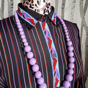 May include: A black button-down shirt featuring vertical stripes in red, purple, and teal, with a geometric pattern on the collar and placket. A long necklace made of smooth, round, light purple beads hangs over the front of the shirt.