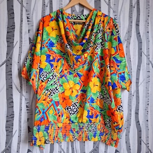 May include: A colourful, short-sleeved blouse with a cowl neck and an elastic waistband. The blouse features a tropical print with yellow, orange, red, green, and blue flowers and geometric patterns. The brand name "La Madina" is visible on the tag.