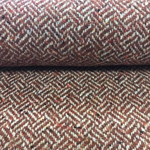 Op de afbeelding: Close-up van een bruine en witte herringbone tweed stof met een subtiele rode vlek. De stof heeft een textuur en een geweven uiterlijk.
