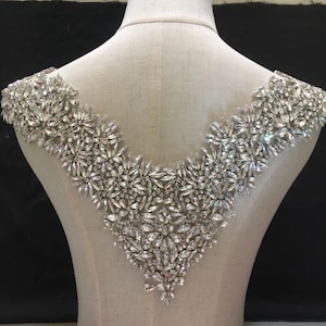 Może przedstawiać: Aplikacja z kryształków w kształcie litery V z przezroczystym wzorem. Aplikacja idealnie nadaje się do dodania blasku sukni ślubnej, sukni wieczorowej lub innej kreacji na specjalne okazje.