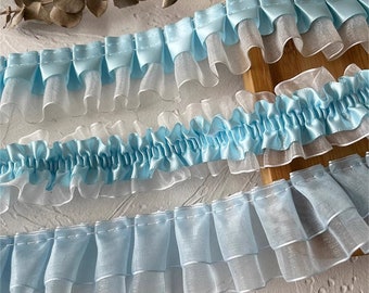 Può includere: Primo piano di tre diversi tipi di bordino blu e bianco con volant. Il bordino è realizzato in un tessuto morbido e trasparente e ha un aspetto delicato e femminile.