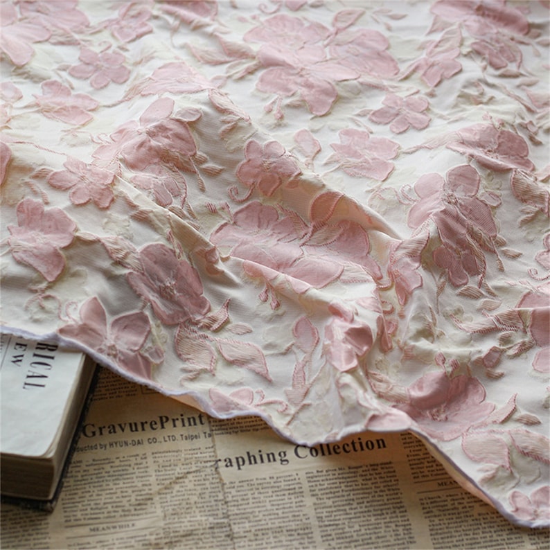 May include: A close-up of a white fabric with a pink floral embossed pattern. The fabric is draped over a newspaper with the text "GravurePrint" and "raphing Collection".