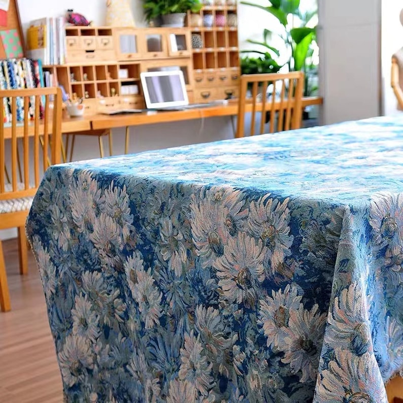 May include: A blue tablecloth with a floral pattern featuring white and pink flowers. The tablecloth is made of a woven fabric and has a slightly textured surface.