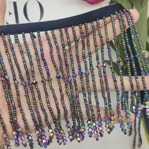 Pode incluir: Close-up de uma guarnição de franja de contas com contas iridescentes em tons de roxo, verde e azul. A franja está presa a uma fita preta.