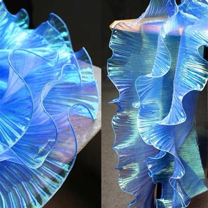 Puede incluir: Una cinta de tela plisada azul iridiscente vibrante con un borde ondulado y con volantes. El material tiene una calidad semitransparente y brillante con propiedades reflectantes que crean un aspecto dinámico y multicolor en los pliegues.