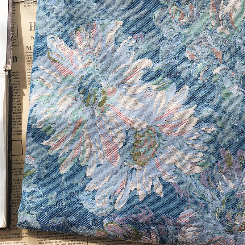 May include: Close-up of a blue fabric with a floral pattern. The flowers are white with pink and green accents. The fabric has a textured woven look.