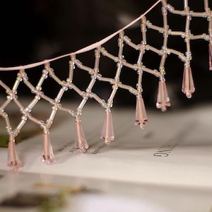 Puede incluir: Primer plano de un delicado adorno de cuentas con una cinta rosa claro. El adorno presenta un patrón de diamantes con pequeñas cuentas transparentes y cuentas rosas en forma de lágrima colgando de la parte inferior. Para manualidades o costura.