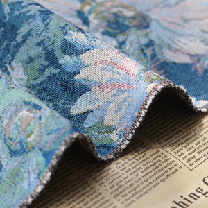 May include: Close-up of a blue and white tapestry fabric with a floral pattern. The fabric has a fringed edge and is folded over a piece of paper with text.