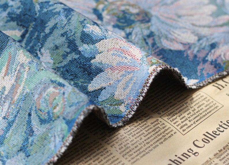 May include: Close-up of a blue and white tapestry fabric with a floral pattern. The fabric has a fringed edge and is folded over a piece of paper with text.