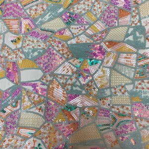 May include: A close-up of a fabric with a geometric pattern made of sequins in shades of pink, teal, gold, and white. The sequins are arranged in a mosaic-like design on a sheer, light-colored fabric.