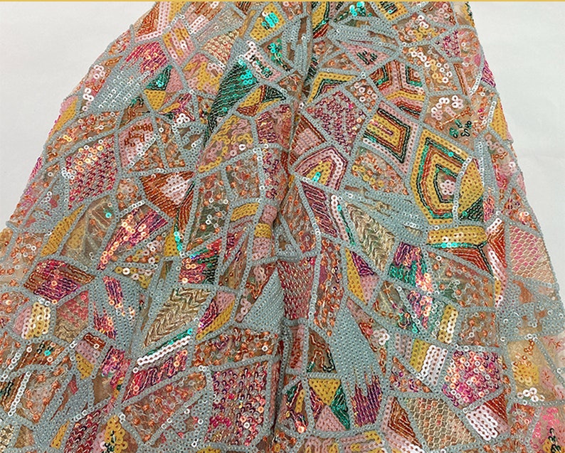 May include: A close-up of a light blue fabric with a geometric pattern of sequins in shades of pink, yellow, green, and orange.