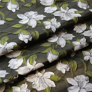 May include: Close-up of a dark green fabric with a white floral pattern. The fabric has a textured, raised design, giving it a three-dimensional look.
