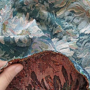 May include: Close-up of a teal and cream floral tapestry fabric with a brown and black woven backing. The fabric is folded over to show the backing.