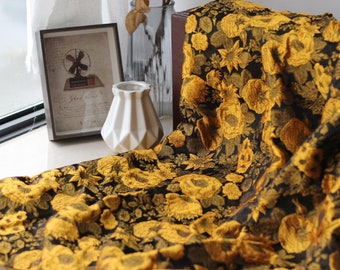 May include: A black fabric with a repeating pattern of large yellow flowers. The fabric is draped, revealing the texture and design. A framed picture, a white vase, and a glass bottle are in the background.