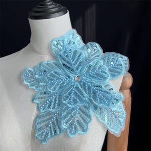 May include: A light blue floral appliqué patch featuring intricate embroidery, shimmering sequins, and small beads. The design includes a central clear rhinestone accent, with layered petals and leaf-like shapes arranged in a large, dimensional flower pattern.