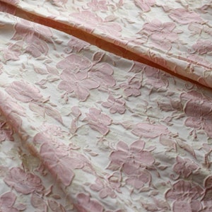 May include: Close-up of a white fabric with a pink floral embossed pattern. The fabric has a textured, raised surface.