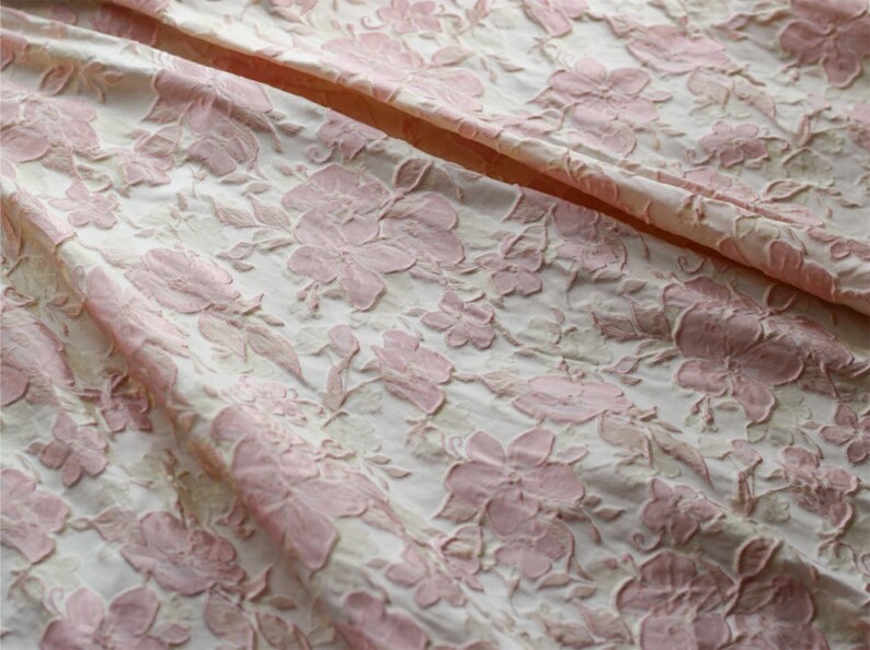 May include: Close-up of a white fabric with a pink floral embossed pattern. The fabric has a textured, raised surface.