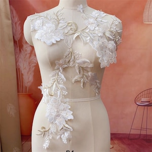 Może przedstawiać: Aplikacja z białej koronki z haftem złotym, z kwiatowymi wzorami i delikatnymi liśćmi. Ta skomplikowana ozdoba idealnie nadaje się do dodania elegancji sukien ślubnych, akcesoriów ślubnych lub innych projektów rękodzielniczych.