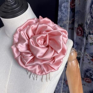 Puede incluir: Un broche de flor grande hecho de tela de satén rosa con múltiples pétalos fruncidos y un acabado suave y brillante. El accesorio está adornado con una serie de pequeñas cuentas blancas similares a perlas que cuelgan de la base.