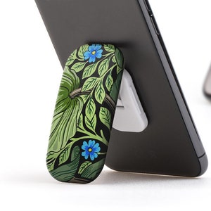 May include: A black smartphone with a floral patterned phone grip stand. The phone is propped up on the stand, and the stand is on a white surface.