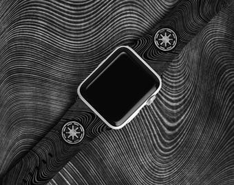 May include: A silver Apple Watch with a black screen and a black and white patterned watch band. The band features two circular embellishments with a starburst design. The watch and band are set against a textured, wavy background.