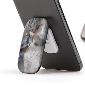 Könnte beinhalten: Zwei schwarze Smartphones mit einem weißen Telefonständer. Der Telefonständer ist ein ausklappbarer Ring mit einem marmorartigen Design.