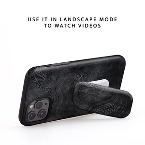 May include: Black phone case with a wood grain pattern and a built-in kickstand. The kickstand is extended and the phone is in landscape mode. "USE IT IN LANDSCAPE MODE TO WATCH VIDEOS" is written in white text on a white background.