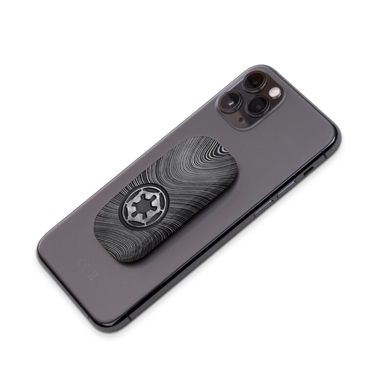May include: A black and silver phone grip featuring a Star Wars Imperial symbol. The grip has a textured, woodgrain-like pattern. It is attached to the back of a dark gray smartphone.