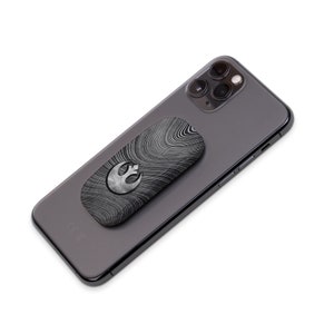 K&ouml;nnte beinhalten: Ein schwarzes Smartphone mit einem silber-schwarzen Handyhalter. Der Halter hat ein Wirbelmuster und ein silbernes Rebellions-Symbol. Das Telefon hat eine Dreifach-Kamera.
