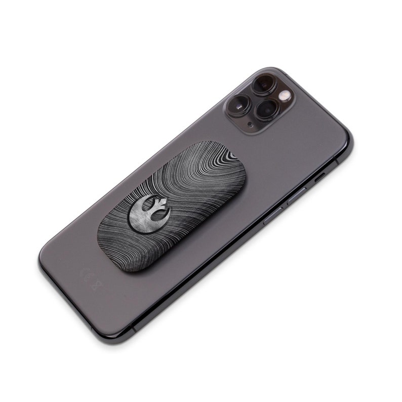 K&ouml;nnte beinhalten: Ein schwarzes Smartphone mit einem silber-schwarzen Handyhalter. Der Halter hat ein Wirbelmuster und ein silbernes Rebellions-Symbol. Das Telefon hat eine Dreifach-Kamera.