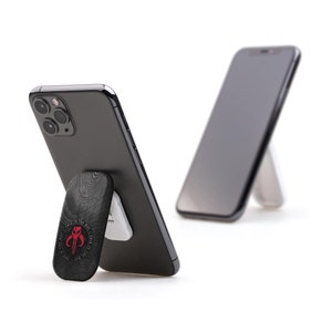 May include: A black smartphone with a pop-out stand featuring a Mandalorian symbol. The stand is made of wood with a dark wood grain pattern. The phone is on a white surface.