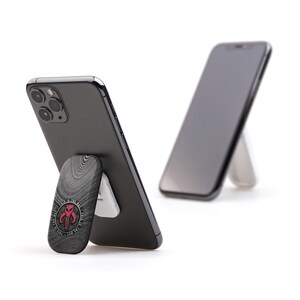 May include: A black smartphone with a textured, gray phone stand featuring a red and white Mandalorian symbol. The phone is propped up in a vertical position.